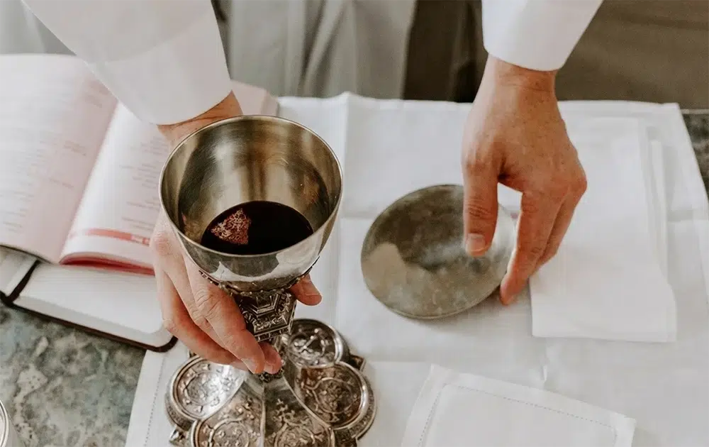 Manos de un sacerdote sosteniendo vasos sagrados de plata, un cáliz labrado con vino y una patena, sobre un altar con un misal abierto durante la misa.