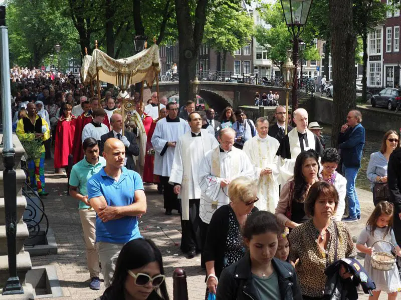 Kerumunan umat beriman mengambil bagian dalam prosesi Corpus Christi di jalan-jalan kota di Eropa, mengiringi Sakramen Mahakudus di bawah kanopi.