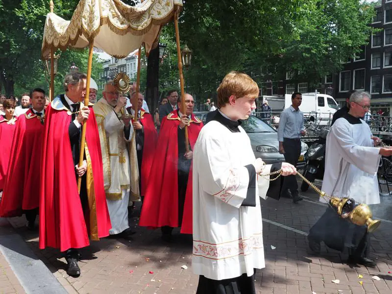 Prosesi Corpus Christi dengan para imam dan umat membawa Sakramen Mahakudus di bawah kanopi di sepanjang jalan perkotaan.