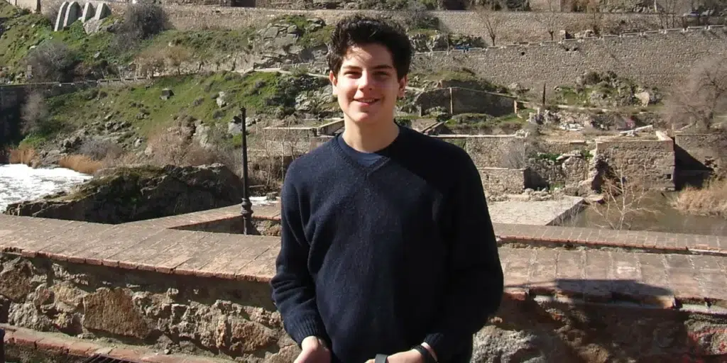 Fotografía del adolescente san Carlo Acutis sonriendo a la cámara en un entorno histórico, con un puente de piedra y un río al fondo, durante un viaje.