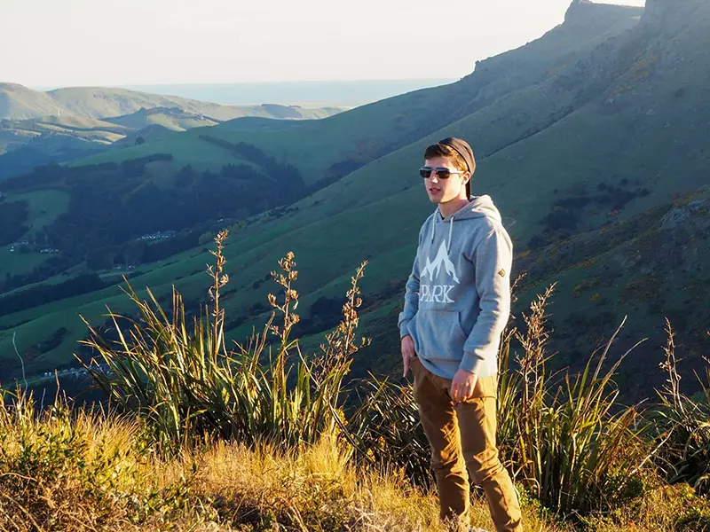 Samuel, joven neozelandés, posa en la cima de una montaña rodeado de naturaleza, con sudadera y gafas de sol.