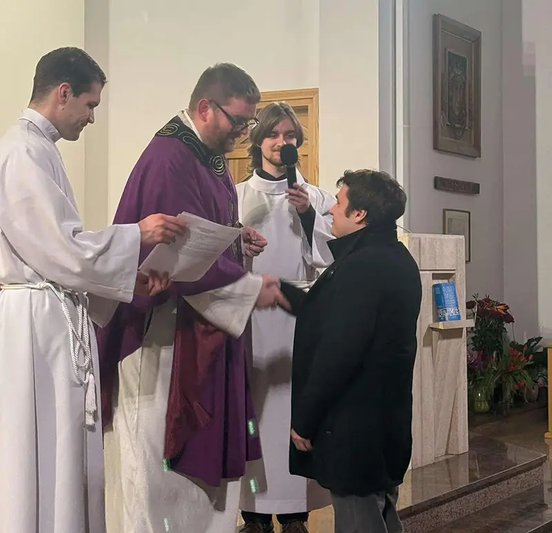 Krištofs Patejuks, poļu priesteris violetā stulā, paspiež roku vīrietim civilajās drēbēs, bet divi citi vīrieši baltās sutanās skatās uz viņu. Viens no viņiem tur mikrofonu. Sižeta darbība notiek baznīcā.