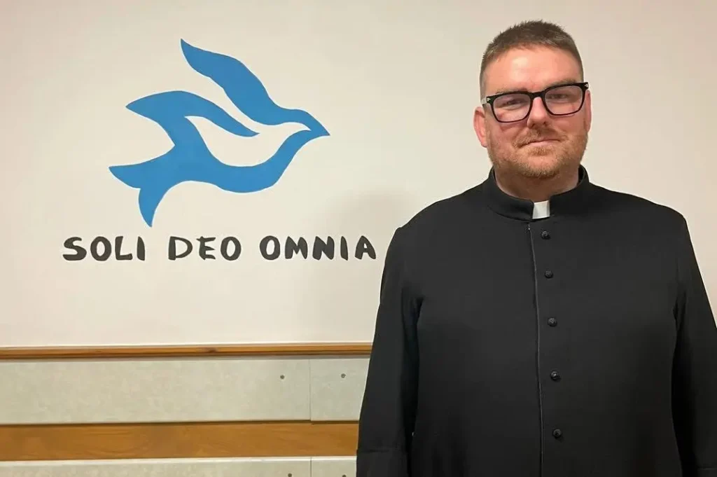 Krištofs Patejuks (Krisztof Patejuk) ar brillēm, tērpies melnā sutanā un romiešu apkakle, stāv baltas sienas priekšā ar zilu balodi un uzrakstu "SOLI DEO OMNIA".