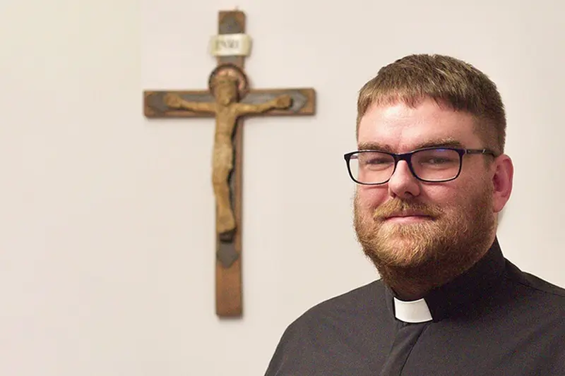 Polish Catholic priest with collar and glasses, standing next to a wooden crucifix hanging on the wall.