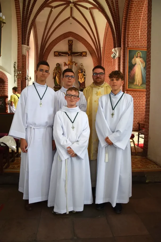Četri bērni un poļu priesteris baltās un zelta liturģiskās drēbēs baznīcā. Bērniem ap kaklu ir krusti, bet priesterim - stula. Fonā redzamas ķieģeļu arkas un altāris ar krucifiksu.