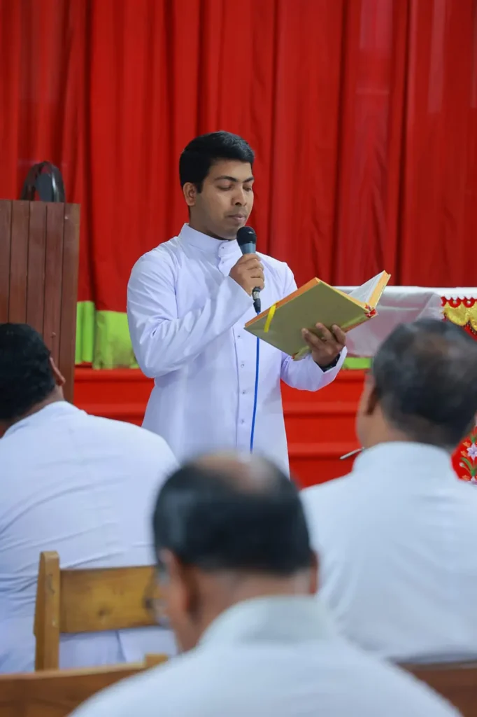 Primer plano del diácono Melwin Thurakal leyendo y hablando por micrófono en un acto relacionado con su ordenación diaconal.
