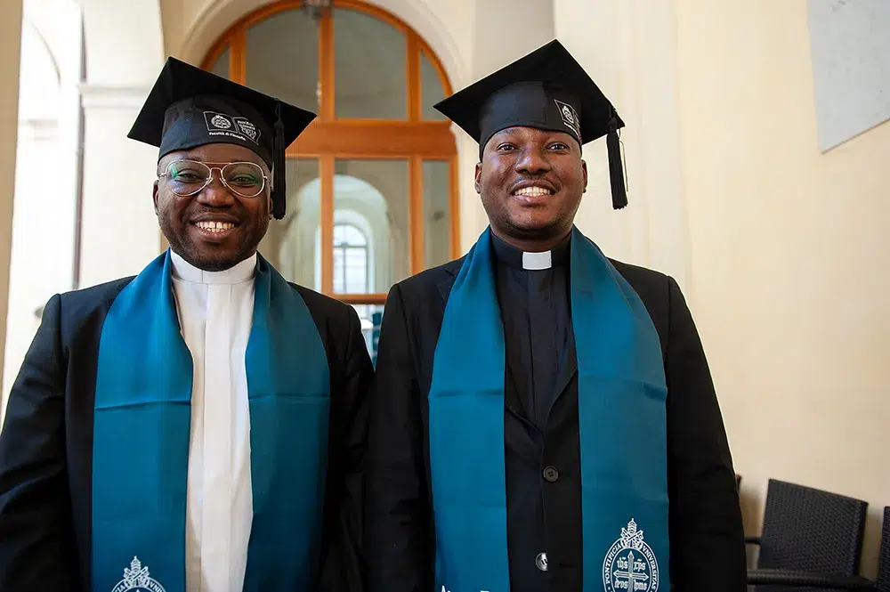 Dos sacerdotes formados en la PUSC, con birrete y estola académica, sonríen tras finalizar sus estudios, ejemplo del impacto de las Herencias destinadas a la formación en la Iglesia.